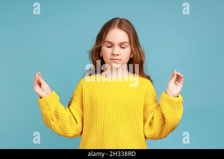 Ritratto di bambina che tiene le braccia in mudra gesto, sensazione di calma rilassato con espressione pacifica, indossando giallo casual stile maglione. Studio interno girato isolato su sfondo blu. Foto Stock