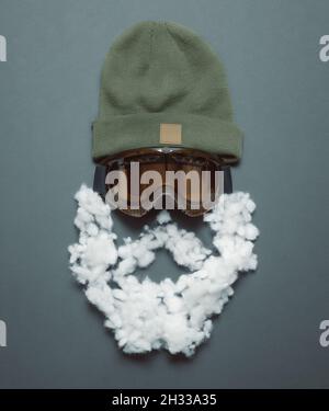 Un cappello sportivo in maglia color palude con una maschera snowboarder e una barba bianca imbottita giacciono su uno sfondo grigio-blu. Vista dall'alto Foto Stock