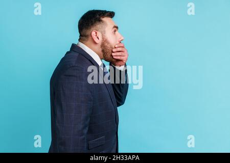 Non dirò nessuno. Vista laterale dell'uomo barbuto che indossa vestito di stile ufficiale che copre la bocca con la mano, mantenendo terribile verità segreta, non vuole parlare. Studio interno girato isolato su sfondo blu. Foto Stock