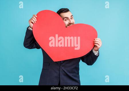 Uomo positivo che indossa un abito ufficiale nascosto dietro un grande cuore rosso e guarda la macchina fotografica con occhi giocosi, tenendo simbolo di amore e affetto. Studio interno girato isolato su sfondo blu. Foto Stock