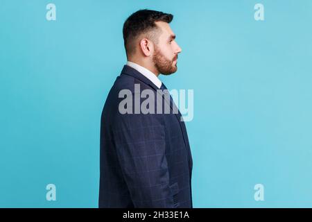 La vista laterale di uomo d'affari bearded che indossa il vestito ufficiale scuro di stile, avendo espressione facciale seria, essendo sconvolto, ha problemi sul lavoro. Studio interno girato isolato su sfondo blu. Foto Stock