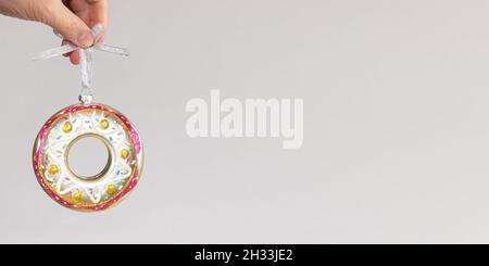 La mano della donna sta tenendo una ciambella colorata di vetro del giocattolo di Natale appesa sul nastro d'argento su sfondo grigio neutro. Creativo minimo Natale largo b Foto Stock