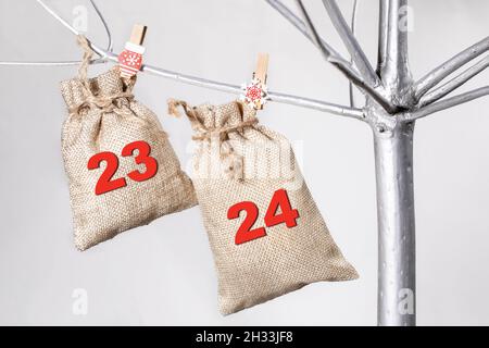 Due borse fatte di matting per regali e numeri 23 e 24 su di loro sono appesi su albero di Natale alternativo fatto di legno dipinto in color argento. Moder Foto Stock
