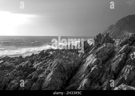 La frastagliata costa rocciosa di Tsitsikamma lungo la costa sudafricana meridionale in bianco e nero, creando un'atmosfera inquietante. Foto Stock