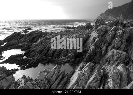 La frastagliata costa rocciosa di Tsitsikamma, con una piscina marea in primo piano, lungo la costa sudafricana meridionale in bianco e nero Foto Stock