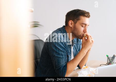 Primo piano di affamato freelance maschio mangiare hamburger guardare video online sul computer portatile durante la pausa. Foto Stock