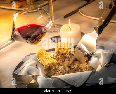 Tartufi bianchi d'Alba in tovagliolo con tagliolini di uova crude e taglierina di tartufo d'acciaio al tavolo del ristorante con candela a legna e calice di vino rosso, neb Foto Stock