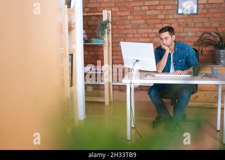 Vista frontale del vecchio programmatore di frellance giovane annoiato che lavora su un computer desktop seduto alla scrivania dell'ufficio di casa, in camera con interni moderni. Foto Stock