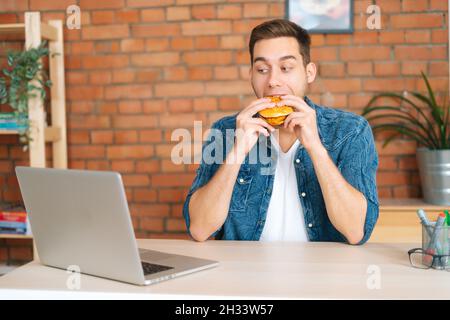 Vista frontale del bel giovane uomo occupato mangiare hamburger guardando il cinema online sul computer portatile seduto alla scrivania in ufficio. Foto Stock