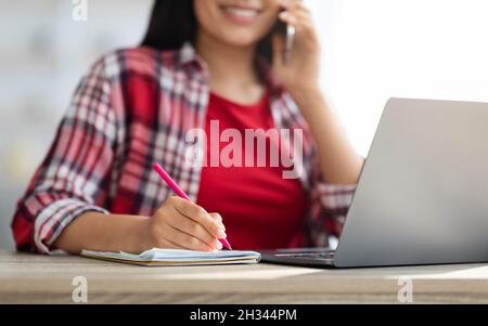 Ufficio domestico. Primo piano Shot di giovane donna parlare sul cellulare e prendere appunti mentre si lavora alla scrivania con computer portatile, irriconoscibile sorridente Free Foto Stock