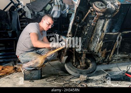 Giovane operaio meccanico che ripara una vecchia carrozzeria di auto d'epoca in un garage disordinato Foto Stock