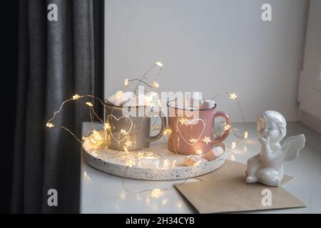 Decorazione appartamento per Natale. Tazze grigie e rosa con cuori bianchi dipinti con cacao, marshmallow su vassoio alla finestra, composizione decorati Foto Stock