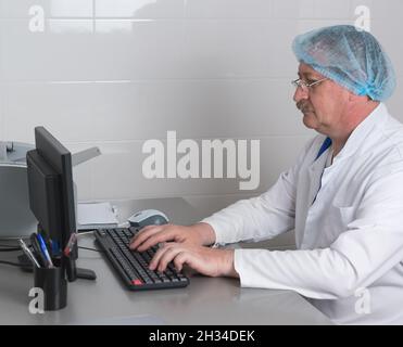 Un medico anziano di sesso maschile in occhiali e vestiti medici è seduto alla sua scrivania in ufficio e digitando rapporto esame sul computer Foto Stock