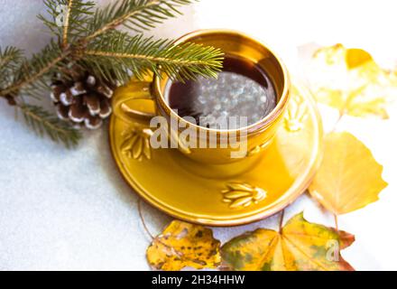 Festa del tè in autunno. Una piccola tazza gialla di caffè in ceramica sul piatto si trova su un tavolo bianco vicino ad un ramo di albero di Natale, cono di abete, foglie cadute. Bellissima Foto Stock