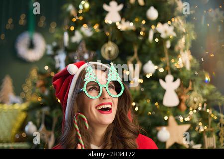 Ora di Natale. Sorridente casalinga di mezza età con cappello di Santa e canna di caramella vicino albero di Natale in casa moderna. Foto Stock