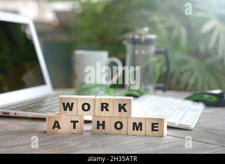 Lavorare a casa, parola scritta su cubi di legno con computer portatile, caffè e piante verdi sullo sfondo. Foto Stock