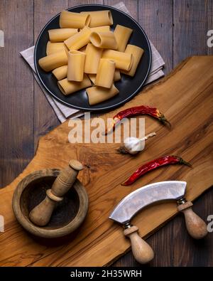 Un tagliere in legno con aglio, peperoncino rosso e pasta cruda con un mortaio in legno e un tritatutto a mezzaluna a doppio manico nelle vicinanze Foto Stock