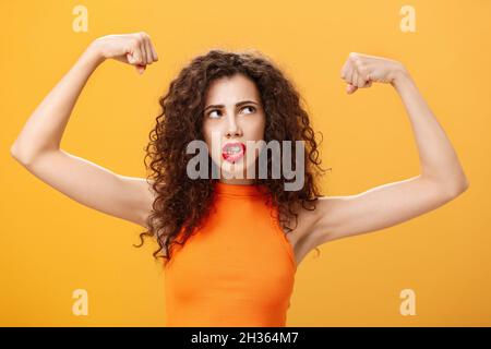 Donna sensazione potente e forte alzare le mani con pugni clenched facendo faccia intensa che si sta allenando in palestra mostrando muscoli e bicipiti guardando Foto Stock
