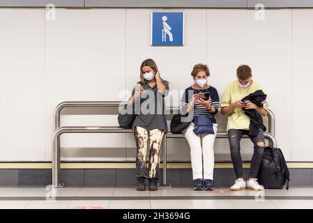Una donna, un uomo e una donna più anziana seduta ad una fermata della metropolitana guardando i loro telefoni cellulari indossare maschera Foto Stock