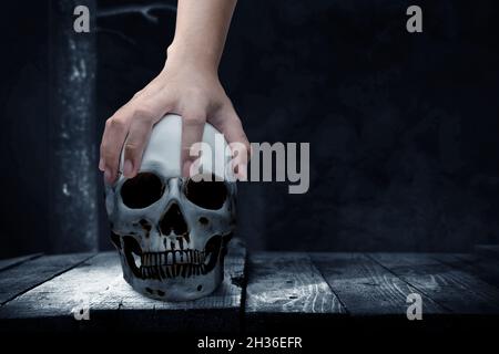 Mano che tiene un cranio umano su un tavolo di legno con sfondo scuro Foto Stock