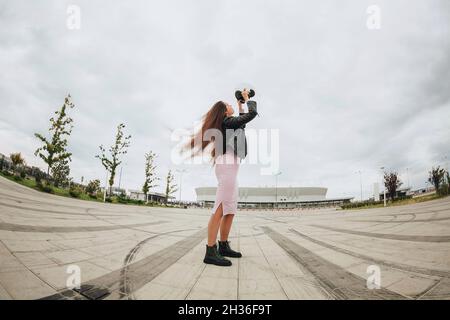 Vista dal basso di giovane donna attraente biker indossando casco di sicurezza per bicicletta mentre si trova in piedi allo stadio vuoto all'aperto, bella motociclista femminile a dres Foto Stock