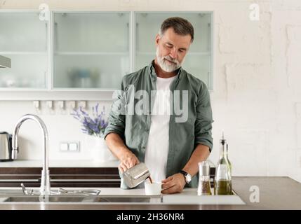 Uomo maturo sorridente che versa un gustoso caffè caldo dalla turca del ferro alla tazza. Foto Stock