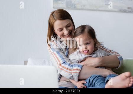 Madre sorridente che abbraccia la figlia con la sindrome di Down che attacca la lingua vicino laptop sfocato Foto Stock