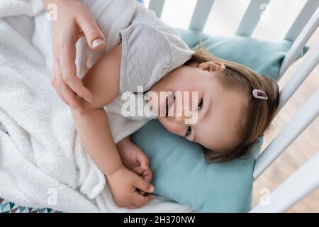Vista dall'alto del bambino positivo con sindrome di Down sdraiato sul presepe vicino a mamma sfocata Foto Stock