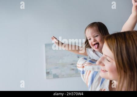 Capretto con sindrome di Down che attacca la lingua vicino alla madre sfocata a casa Foto Stock