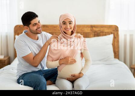 Giovane musulmano amorevole che si aspetta coppia coccolare mentre si passa il tempo insieme, uomo massaggiando le spalle della moglie incinta Foto Stock