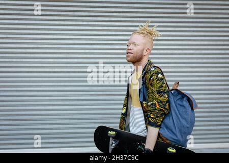 Felice albino afroamericano uomo con dreadlock camminare tenendo skateboard Foto Stock