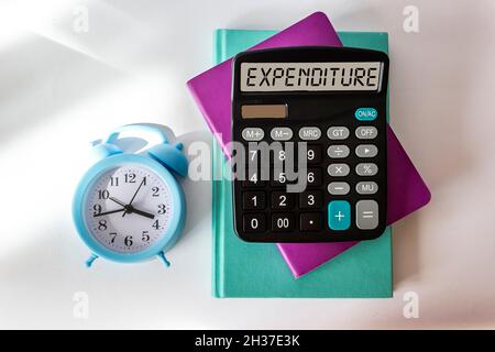 Calcolatrice con la parola SPESA sul display della calcolatrice. I notepad e la sveglia si trovano su uno sfondo bianco. Foto Stock