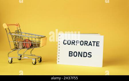 Blocco note con il testo Corporate Bonds su sfondo giallo. Carrello giocattolo con monete Foto Stock