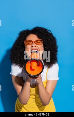 eccitata donna afroamericana in occhiali arancioni urlando in megaphone su blu Foto Stock