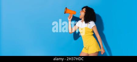 energica donna afroamericana in tuta trendy urlando in megaphone su blu, banner Foto Stock