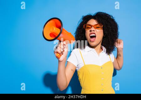 eccitata donna afroamericana in occhiali che gridano in arancione altoparlante su blu Foto Stock