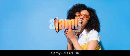 eccitata donna afroamericana in occhiali gridando in altoparlante su blu, banner Foto Stock