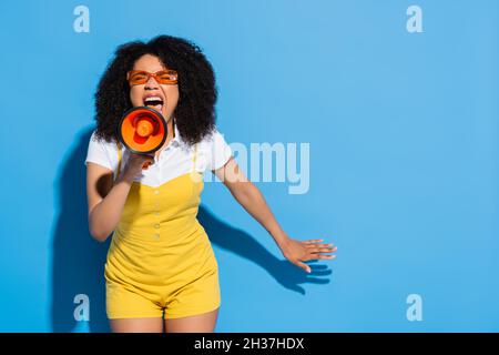 donna afroamericana in occhiali arancioni urlando in megaphone su blu Foto Stock