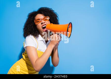 agitato afroamericana donna in occhiali trendy urlando in megaphone su blu Foto Stock