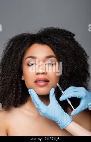 estetista in guanti di lattice che danno l'iniezione di bellezza alla donna afroamericana isolata su grigio Foto Stock