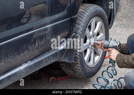 L'operatore che lavora con una chiave pneumatica svitate i bulloni dalla ruota dell'auto. Sostituzione della ruota sul centro di assistenza pneumatici. Macchina nera sporca sollevata sulla ja Foto Stock
