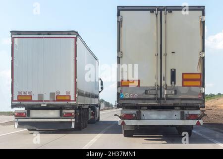 Vista posteriore di due camion di scarico guida fuori della città su una strada asfaltata piatta tra campi contro un cielo blu con le nuvole in una giornata di sole. La strada lavora su un in Foto Stock