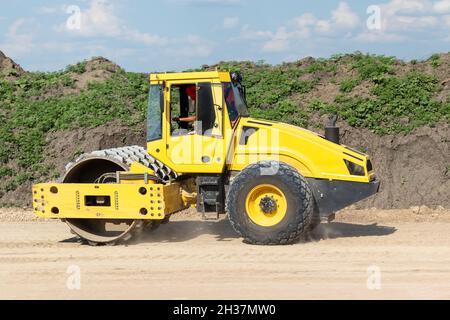 Rullo giallo per la costipazione di superfici morbide nel cantiere fuori città. La strada lavora su un'autostrada interurbana in una giornata nuvolosa Foto Stock