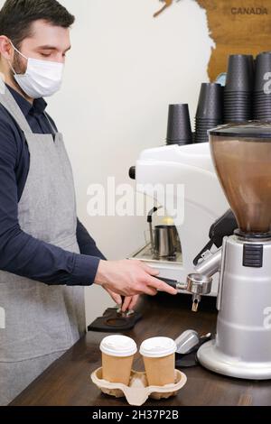 Barista che serve caffè in tazzine da asporto in coffee shop in maschera protettiva. Caffè da gustare durante il coronavirus. Tazze monouso. Macinare il caffè Foto Stock