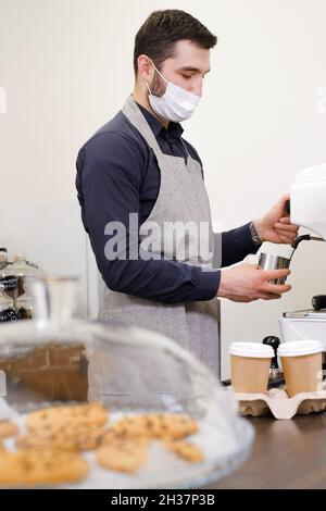 Barista che serve caffè in tazzine da asporto in coffee shop in maschera protettiva. Caffè da gustare durante il coronavirus. Tazze monouso. Macinare il caffè Foto Stock