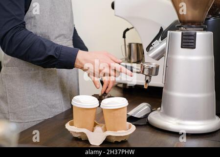 Barista che serve caffè in tazzine da asporto in coffee shop in maschera protettiva. Caffè da gustare durante il coronavirus. Tazze monouso. Macinare il caffè Foto Stock