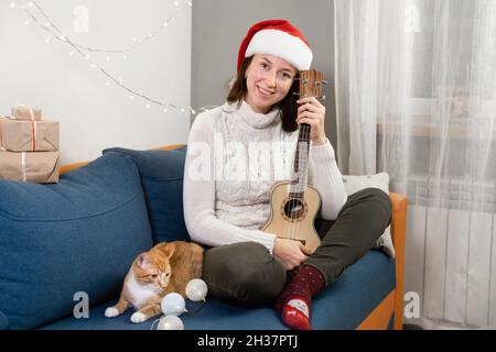 Donna che festeggia il Natale da sola a casa, suonando ukulele (piccola chitarra). Stile di vita felice di festa, sfondo di luci natalizie e. Foto Stock