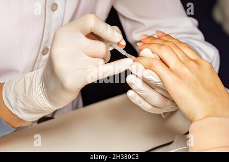 Primo piano di una mano bianca femminile durante una sessione di manicure. Un estetista in guanti di lattice bianco applica smalto incolore unghie ad una giovane donna. Foto Stock