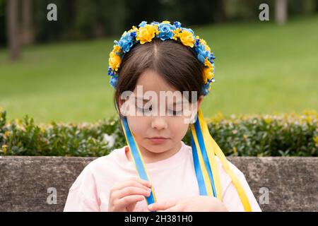 Ragazza triste con decorazione popolare Ucraina sulla sua testa. Il bambino tocca i nastri della corona mentre si siede su una panca all'aperto Foto Stock