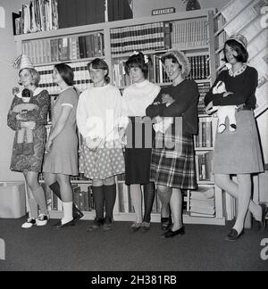 1960 studenti storici, giovani donne delle scuole superiori in una stanza di studio o sezione di una biblioteca in piedi in una linea, con tre di loro che tengono i giocattoli morbidi, con una ragazza sulla sinistra che indossa un cappello da festa e che tiene una bambola del batman, Virginia, USA. Forse partecipare ad una certa specie di classe sul gioco immaginativo. Dietro di loro, in cima a una libreria piena di libri di testo, un segno che dice PENSARE. Foto Stock